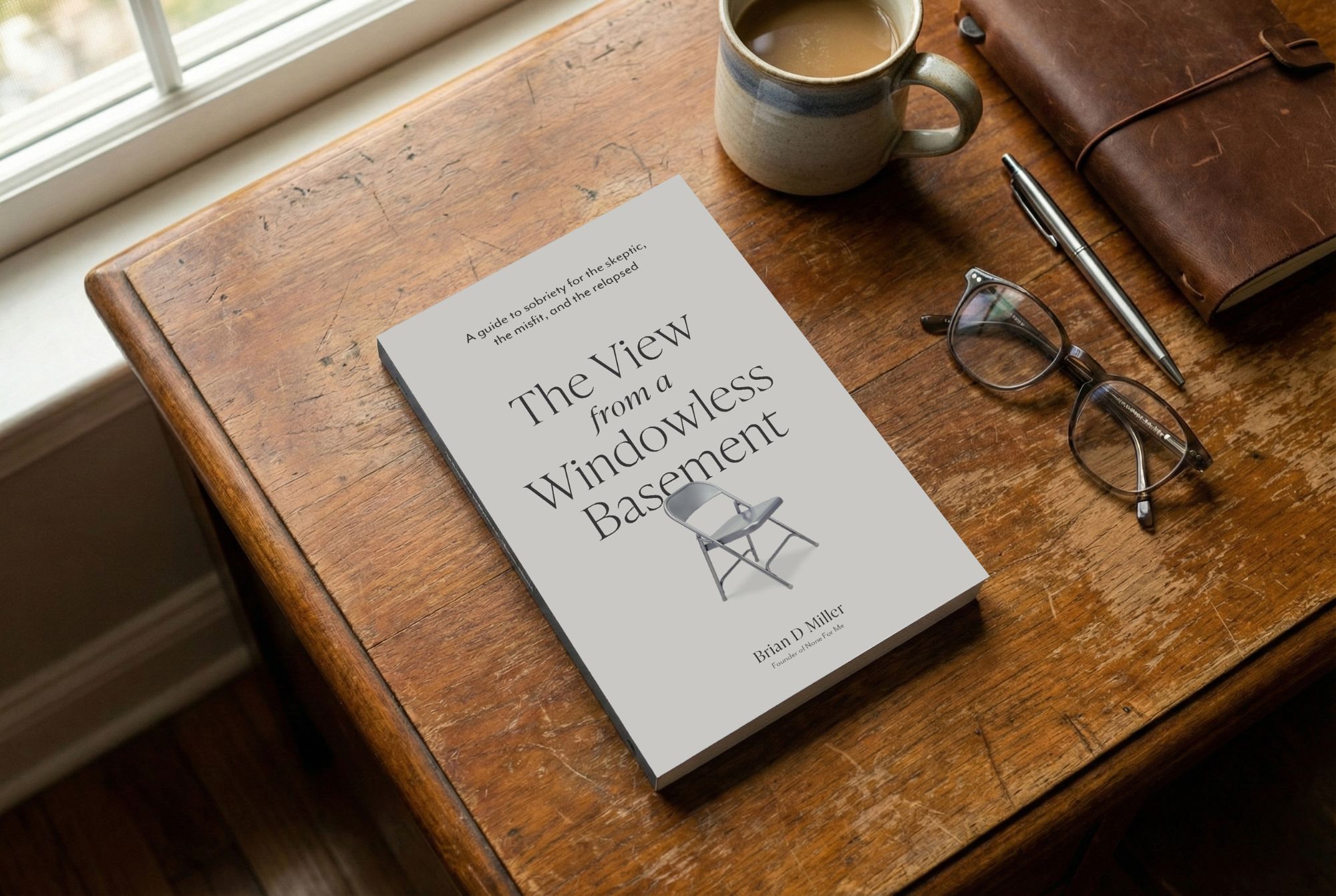 The View from a Windowless Basement resting on a wooden desk by a window, beside a cup of coffee, reading glasses, a pen, and a leather journal.